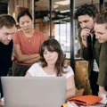 Four colleagues crowd around one desk as a teammate drives a process on her laptop.